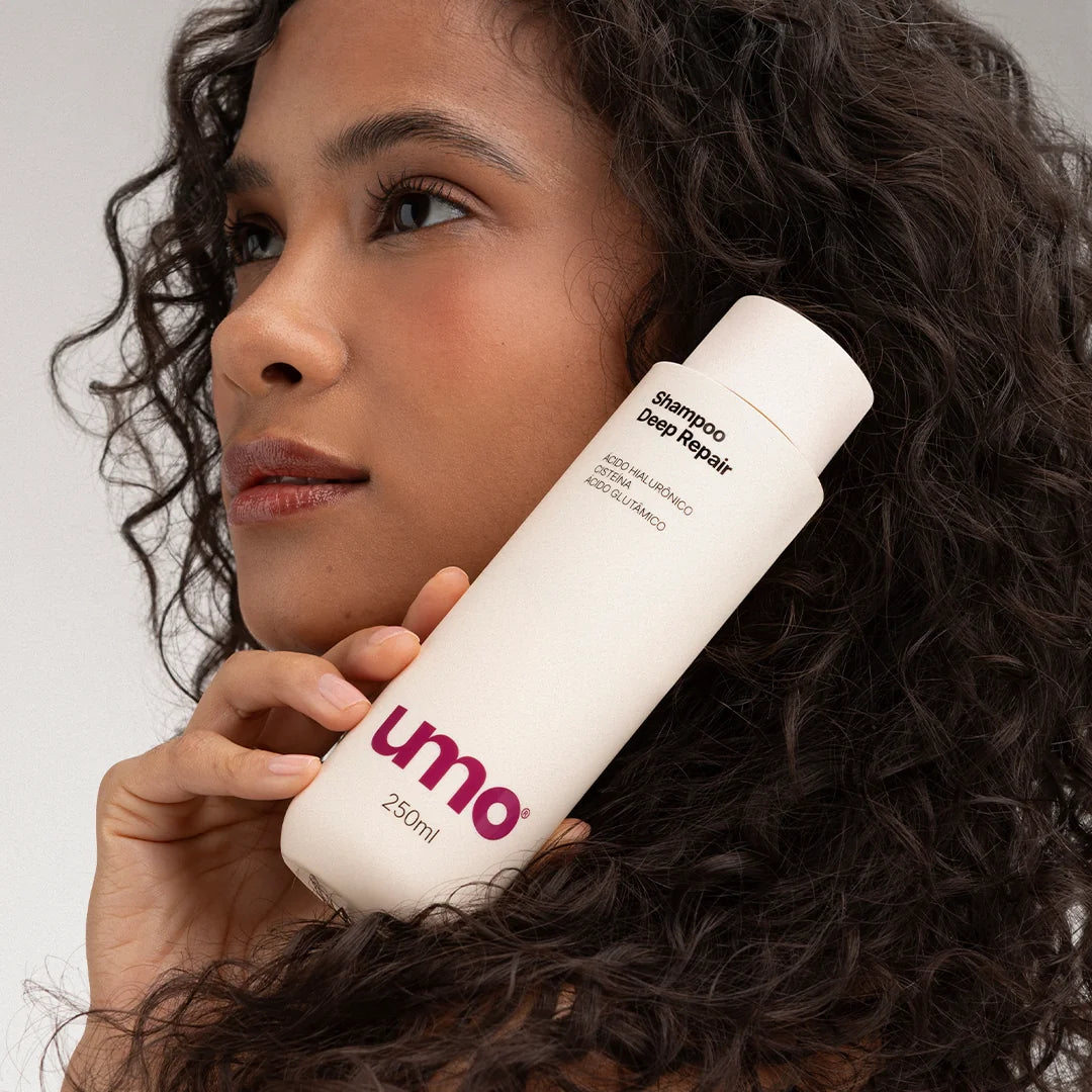 Modelo de cabelo cacheado segurando o Shampoo Deep Repair Umo 250ml ao lado do rosto
